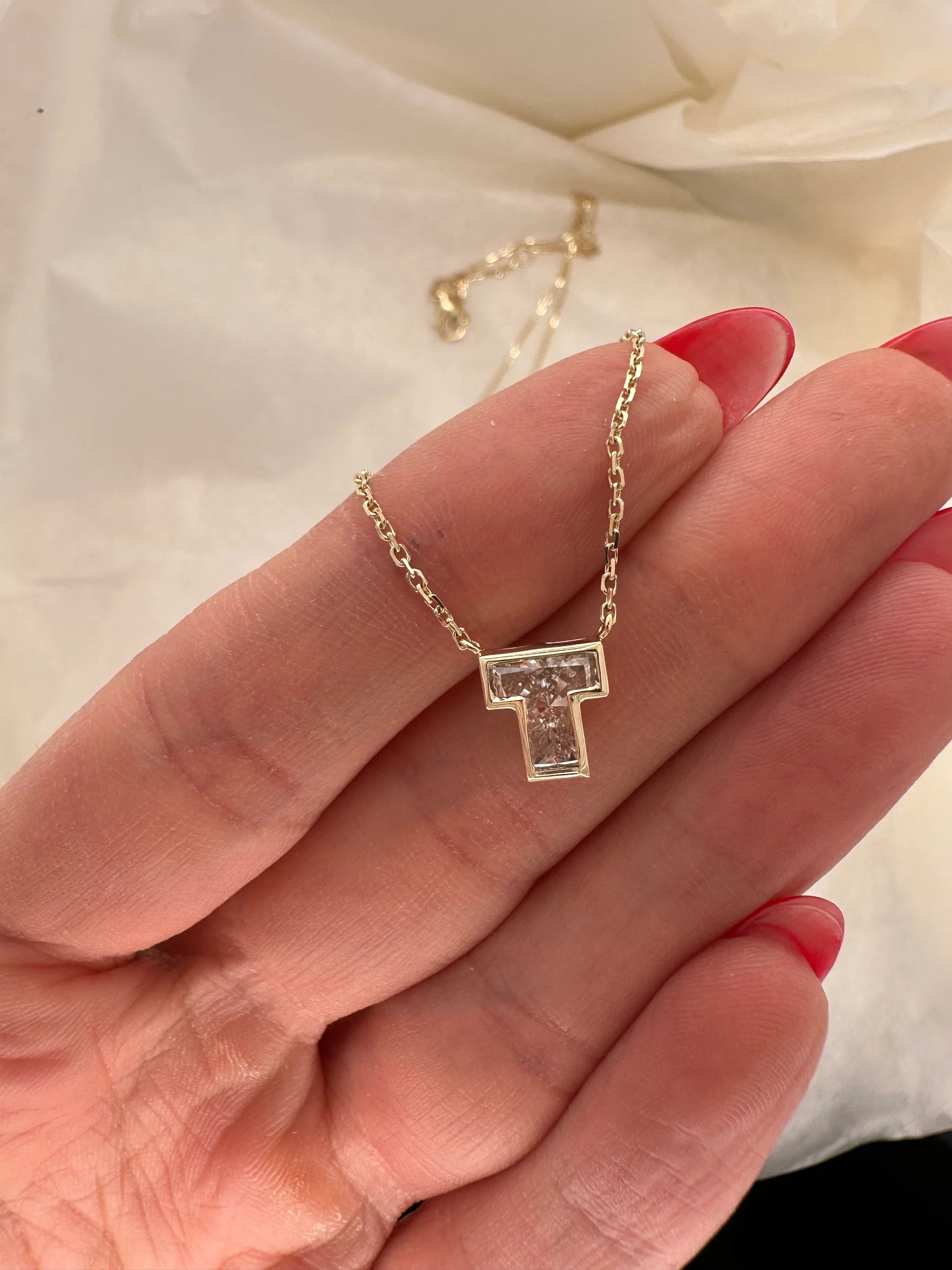 Gold necklace with a 'T' pendant held between fingers on a white fabric background